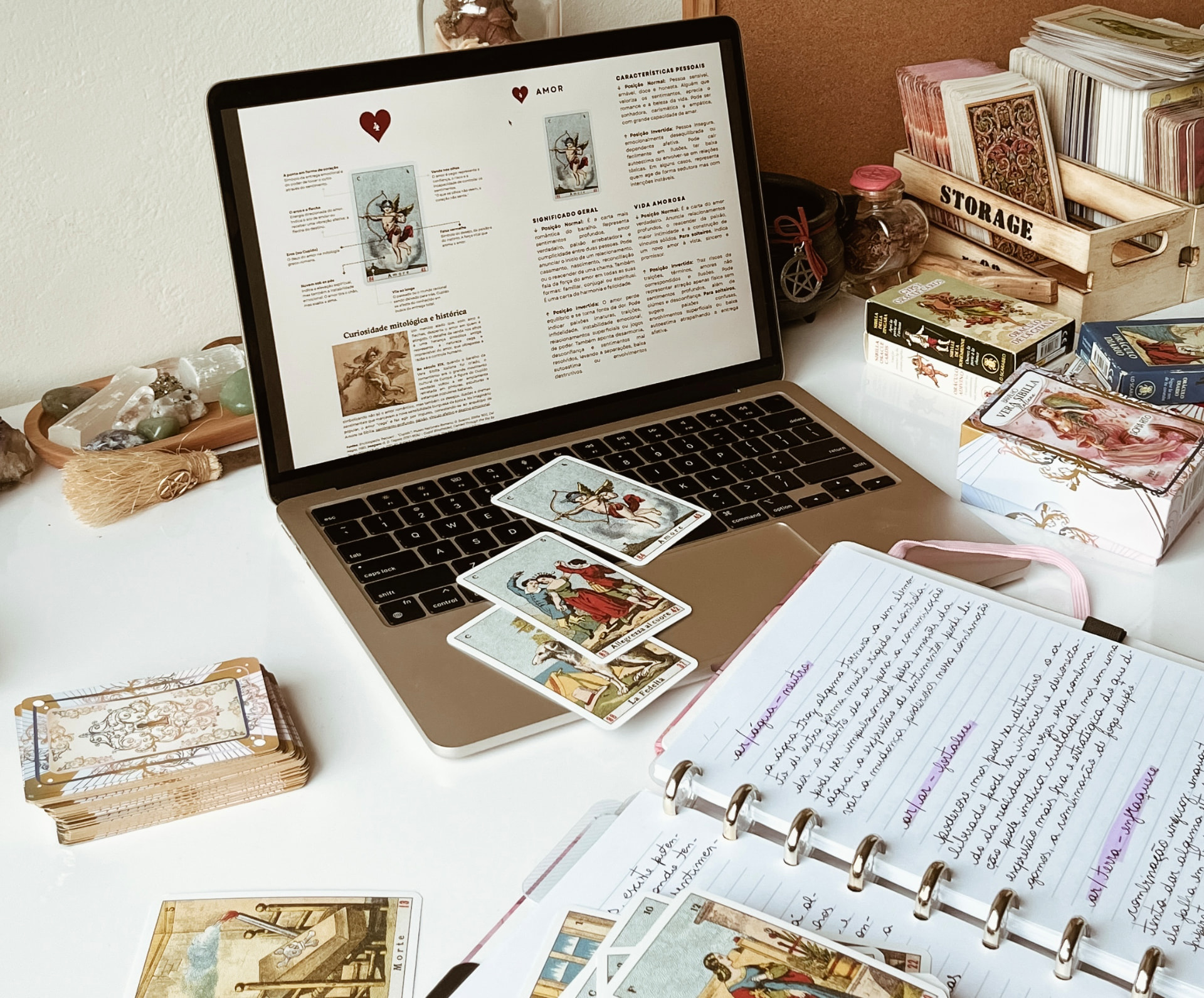 Mesa de estudo com notebook aberto no guia da Vera Sibilla, cartas Amor e outras espalhadas sobre o teclado
