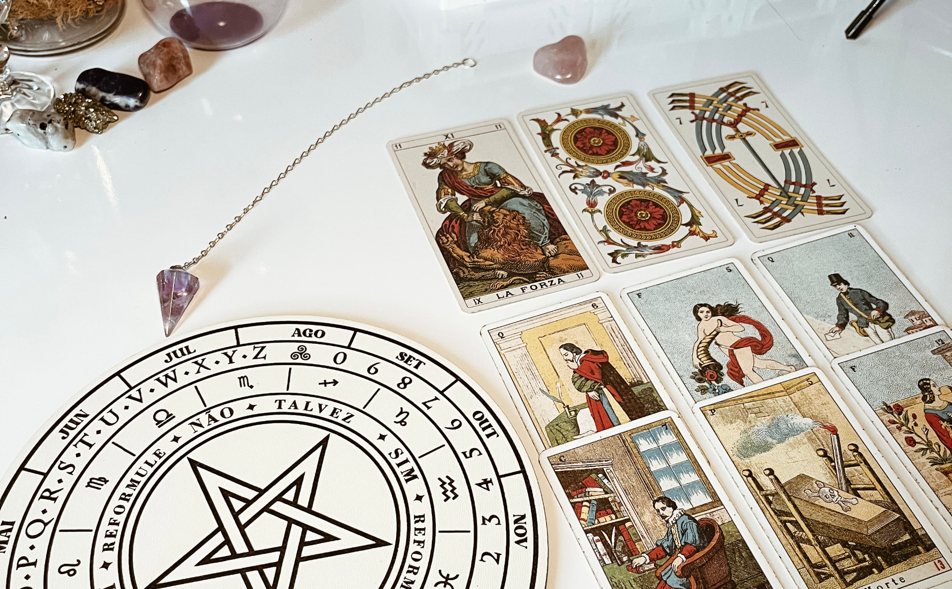 Mesa com tabuleiro de pêndulo, pêndulo de ametista e cartas de Tarot Marselha abertas, incluindo A Força (XI)